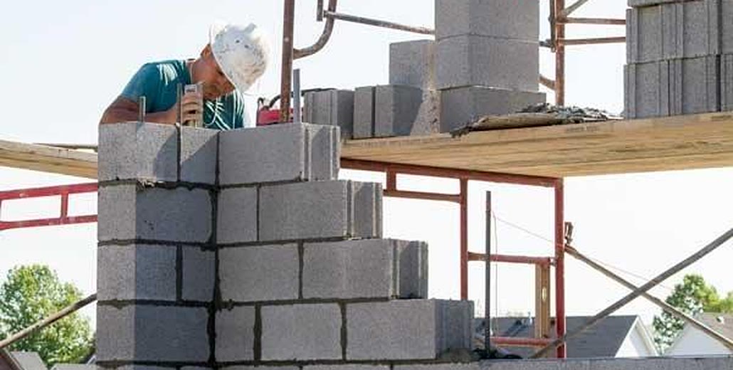 A mason laying concrete block at the corner of a residential foundation wall, with more block stacked on scaffold planks behind him