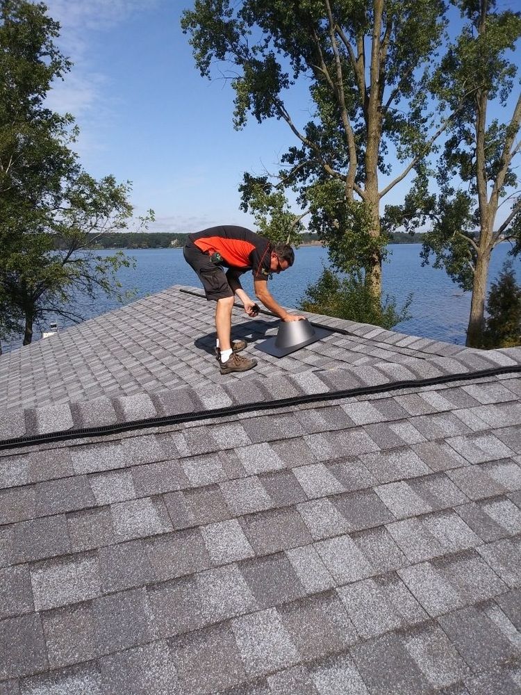 A member of the crew fitting a vent cap on a roof, with the lakeshore behind