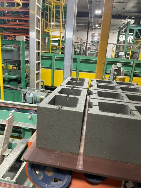 Freshly molded two-core 8 inch concrete blocks sitting on a steel pallet as they come off the block machine, with green plant equipment behind them