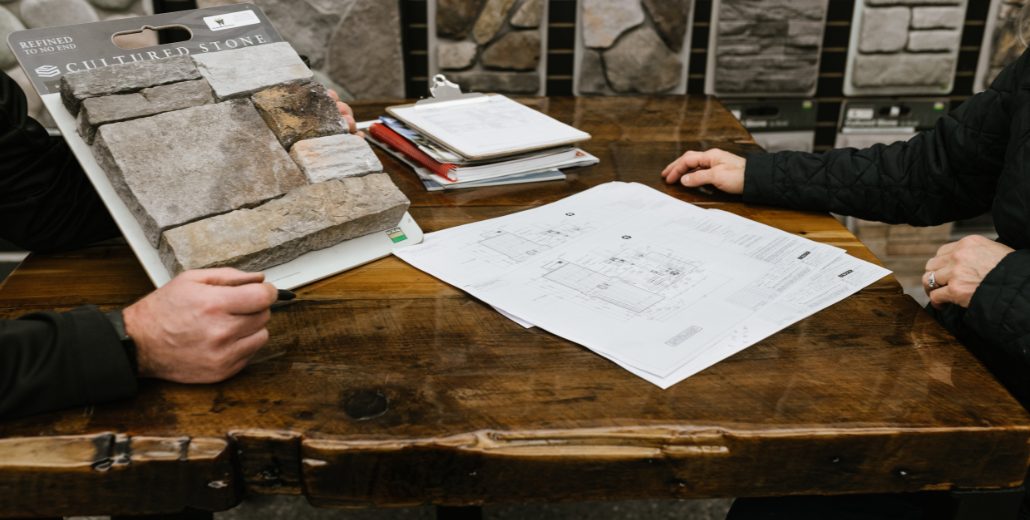 Two people at a wooden showroom table reviewing a Cultured Stone sample board beside a set of construction plans, with stone display panels on the wall behind them