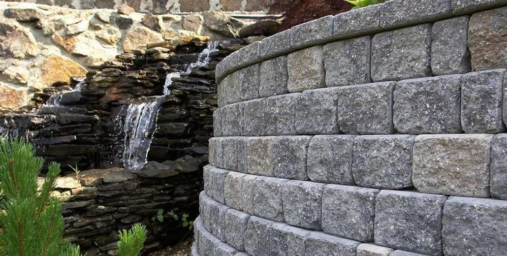 A curved segmental block retaining wall with a capped top course, holding back a planted bank beside a stacked stone water feature