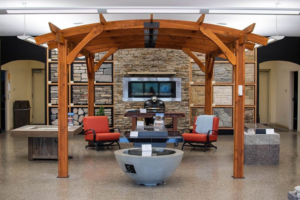 The Spring Lake showroom floor, with stone sample walls, a linear gas fireplace and fire pit tables under a cedar pergola
