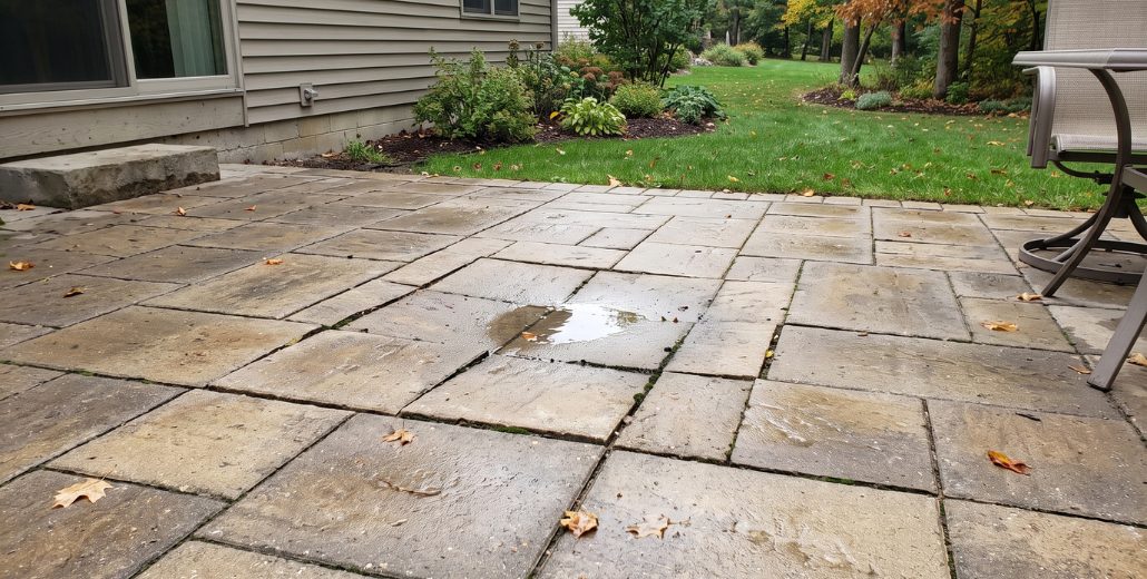 A wet backyard paver patio beside a house, with a shallow puddle sitting in a low spot in the middle of the pavers and fallen leaves scattered across the surface